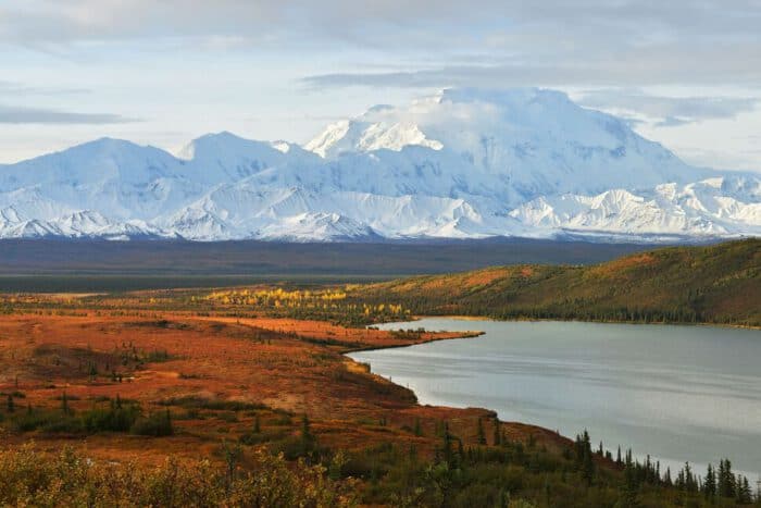 A calm lake bordered by autumn foliage in the foreground with snow-covered mountains in the background under a mostly cloudy sky.