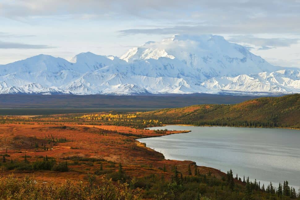 A calm lake bordered by autumn foliage in the foreground with snow-covered mountains in the background under a mostly cloudy sky.