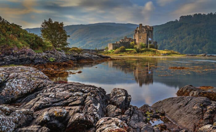 A stone castle sits beside a calm lake, surrounded by rocky shore and green hills under a cloudy sky.