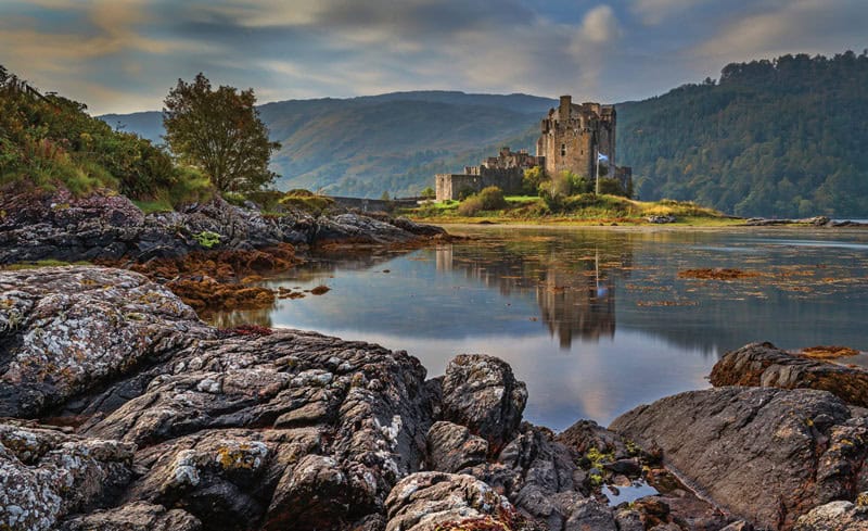 A stone castle sits beside a calm lake, surrounded by rocky shore and green hills under a cloudy sky.