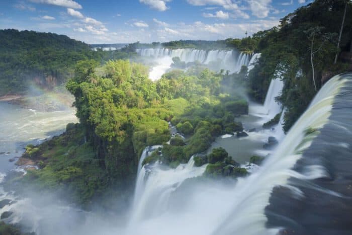 Water cascades from multiple waterfalls over lush green cliffs, surrounded by dense forest and a partly cloudy sky.