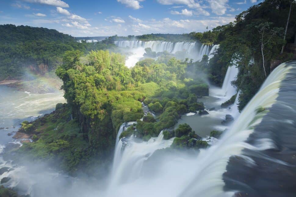 Water cascades from multiple waterfalls over lush green cliffs, surrounded by dense forest and a partly cloudy sky.
