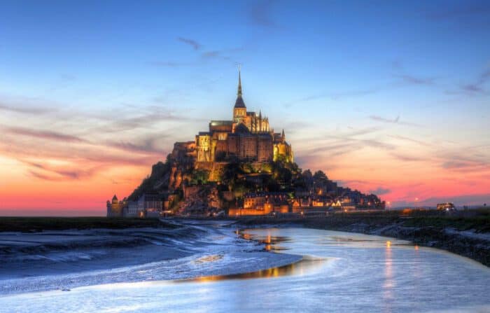 A medieval abbey and village sit atop Mont Saint-Michel island at sunset, with a tidal river in the foreground.