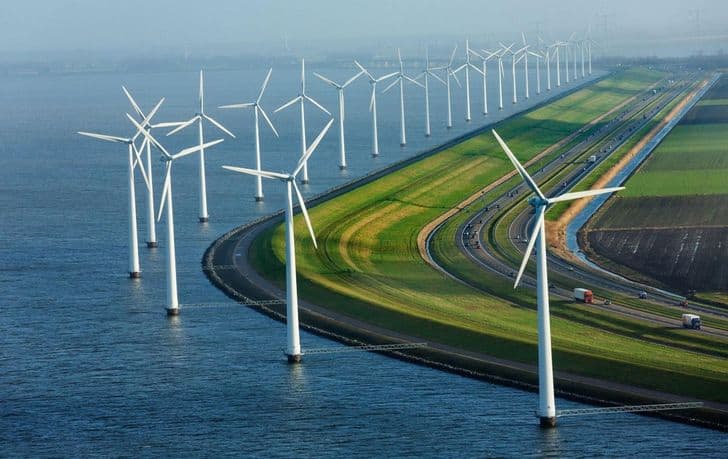 Rows of wind turbines line a coastal road, with water on one side and fields on the other, under a hazy sky.