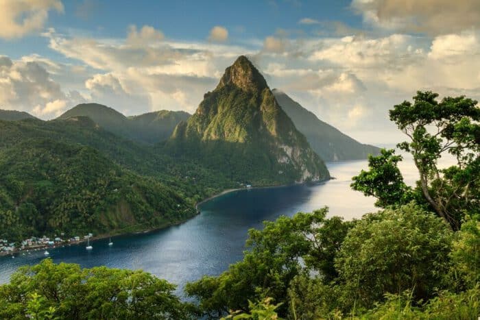 A tall, green mountain rises above the coastline, surrounded by dense forest and a calm blue sea under a partly cloudy sky.