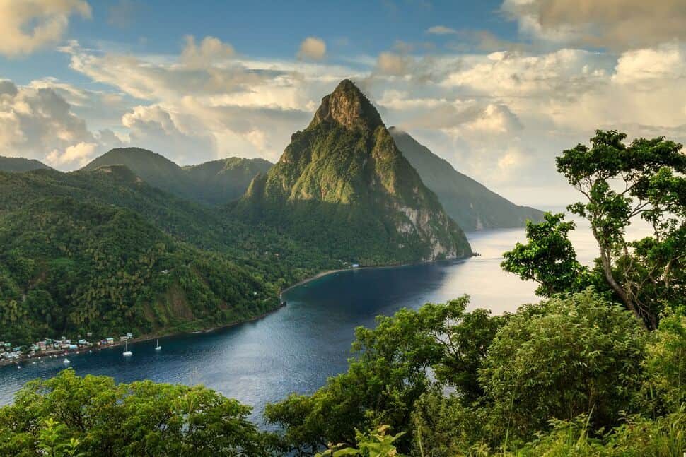 A tall, green mountain rises above the coastline, surrounded by dense forest and a calm blue sea under a partly cloudy sky.