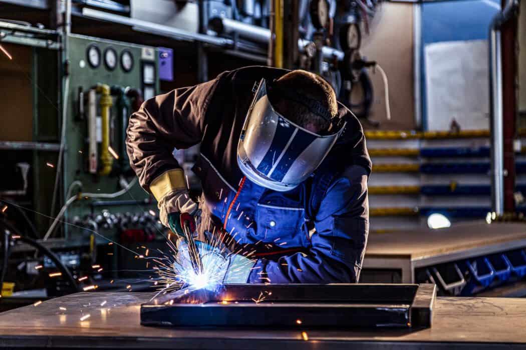 A person wearing protective gear uses a welding torch, creating sparks in an industrial workshop setting.