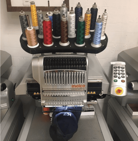 A computerized embroidery machine with multiple colored threads loaded and a dark cap being embroidered underneath the needle.