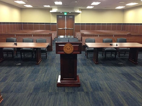 Empty courtroom with a wooden podium in the center, tables and chairs on each side, and rows of benches in the background.