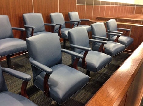 A row of empty blue upholstered chairs with wooden armrests arranged in a jury box inside a courtroom.