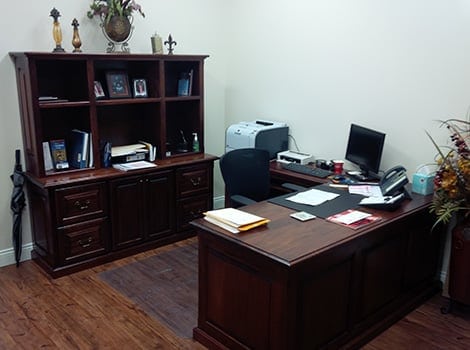 An office with a wooden desk, hutch with shelves, computer, office chair, printer, phone, and various papers and decorations.