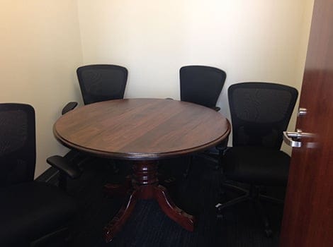 A small meeting room with a round wooden table and four black office chairs, seen through a partially open door.