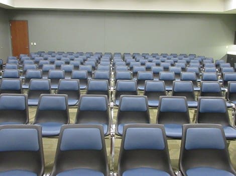 Rows of empty blue and gray chairs are arranged in a conference or meeting room with a closed door on the left.