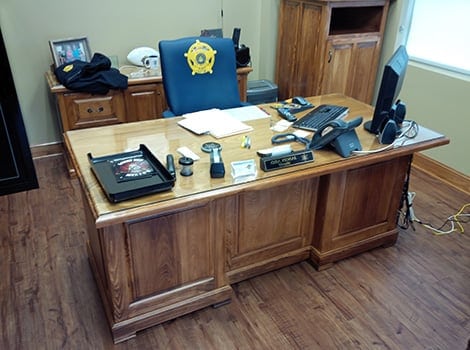 A neatly organized wooden office desk with a sheriff's badge on the chair, paperwork, a phone, computer monitor, and office supplies.