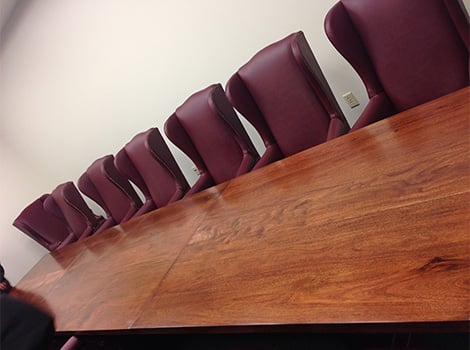 A long wooden conference table with several empty maroon chairs lined up against a plain white wall.