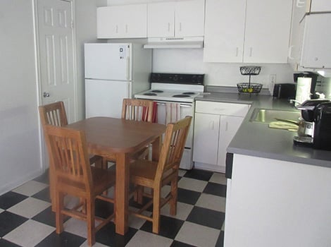 A small kitchen with white cabinets, a square wooden table with four chairs, and a black-and-white checkered floor.