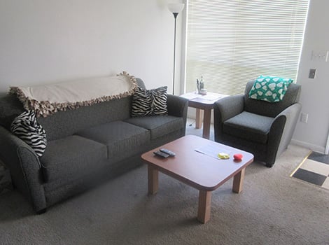 A small living room with a gray couch, an armchair, a wooden coffee table, a lamp, and a window with closed blinds.