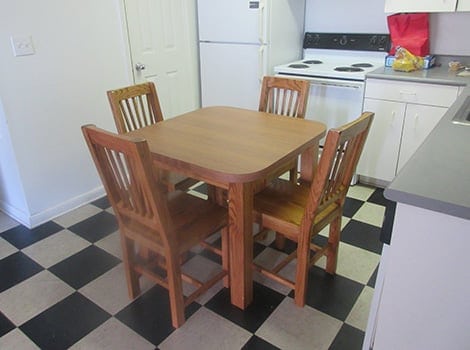 A wooden table with four matching chairs sits on a black and white checkered kitchen floor near white cabinets and appliances.