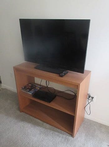 A flat-screen TV sits on a wooden stand with a remote, magazines, and electronic device; cords are plugged into a wall outlet.