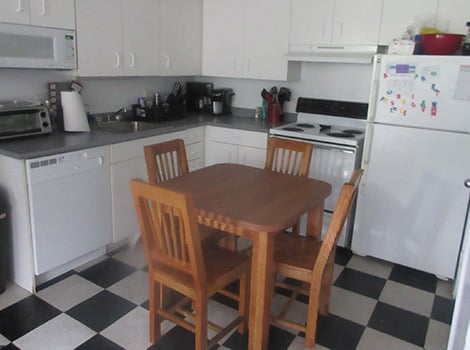 A kitchen with white cabinets, black countertop, white appliances, and a wooden table with four chairs on a black-and-white checkered floor.