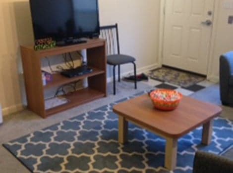 A living room with a TV on a wooden stand, a chair, a small table with a bowl, and a patterned rug on the floor.