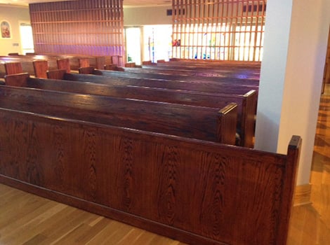 Rows of empty wooden pews are arranged inside a building with light-colored walls and wood flooring.