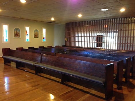 Rows of empty wooden pews in a chapel with wood flooring, stained glass windows, and a decorative wooden wall at the front.