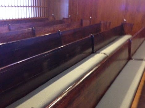 Rows of empty wooden pews with padded seats are arranged in a room with wooden walls and a window with vertical bars.