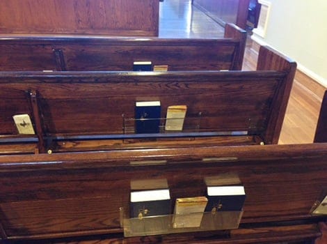 Rows of empty wooden church pews with hymnals and Bibles in holders attached to the backs of the seats.