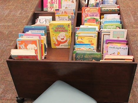 A wooden book cart filled with colorful children's books, next to a light blue cushion on a carpeted floor.