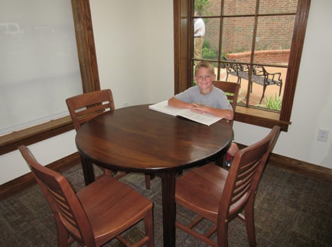 A boy sits at a round wooden table with four chairs, reading a large book near windows in a room with beige walls.