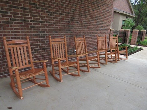 Six wooden rocking chairs are lined up on a concrete porch in front of a brick wall, with a garden and fence visible nearby.