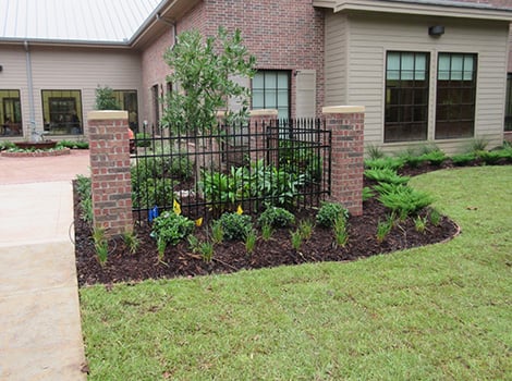 A small garden bed with green plants is bordered by mulch, brick pillars, and a black metal fence next to a brick and siding building.