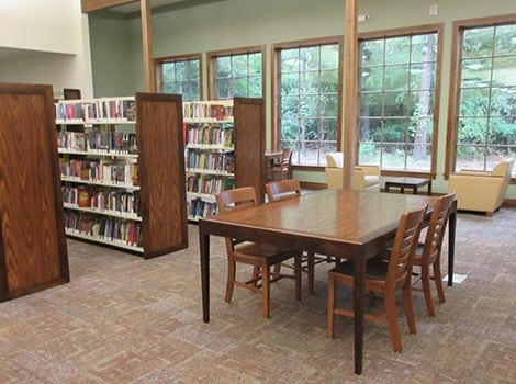 A library room with bookshelves, a wooden table with chairs, armchairs by large windows, and a view of trees outside.