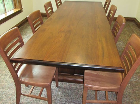 A rectangular wooden table with eight matching wooden chairs in a carpeted room with cream-colored walls and a window.