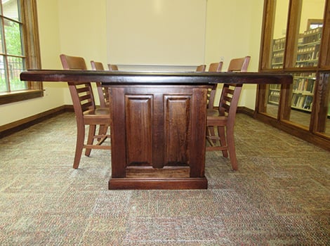 A wooden table with several chairs around it is placed in a carpeted room with windows and bookshelves.