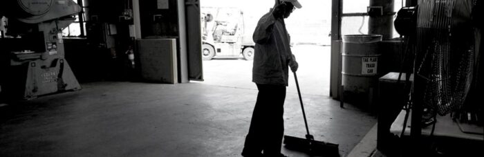 A person wearing a cap sweeps the floor inside a dimly lit industrial garage; sunlight streams in from an open door.