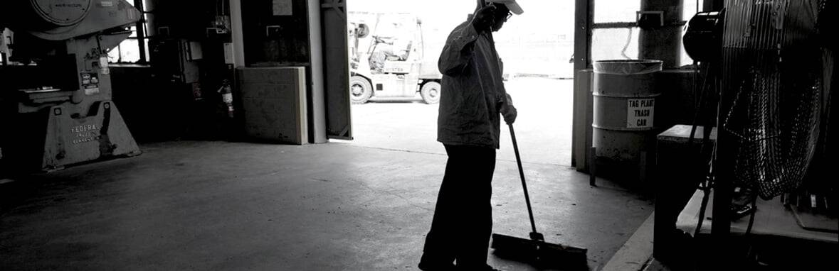 A person wearing a cap sweeps the floor inside a dimly lit industrial garage; sunlight streams in from an open door.