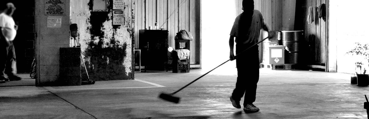 A person sweeps the floor of an industrial warehouse with a large broom; another person stands in the background near a wall.