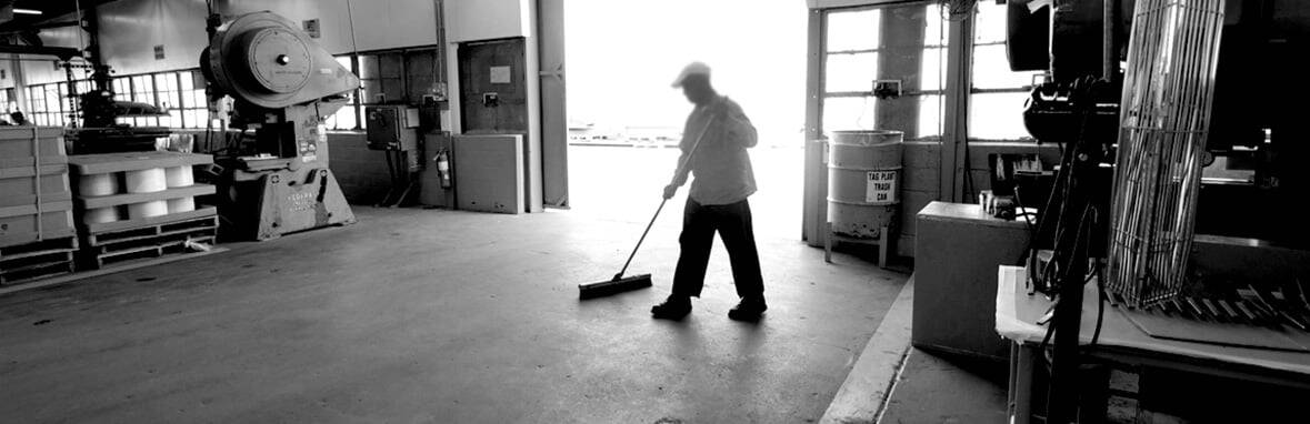 A person sweeps the floor inside a spacious industrial workshop with large open doors and various equipment in view.