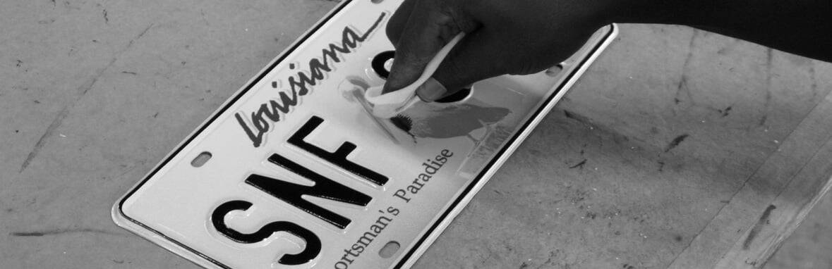 A hand wipes a newly made Louisiana license plate with the letters "SNF" on a work surface.