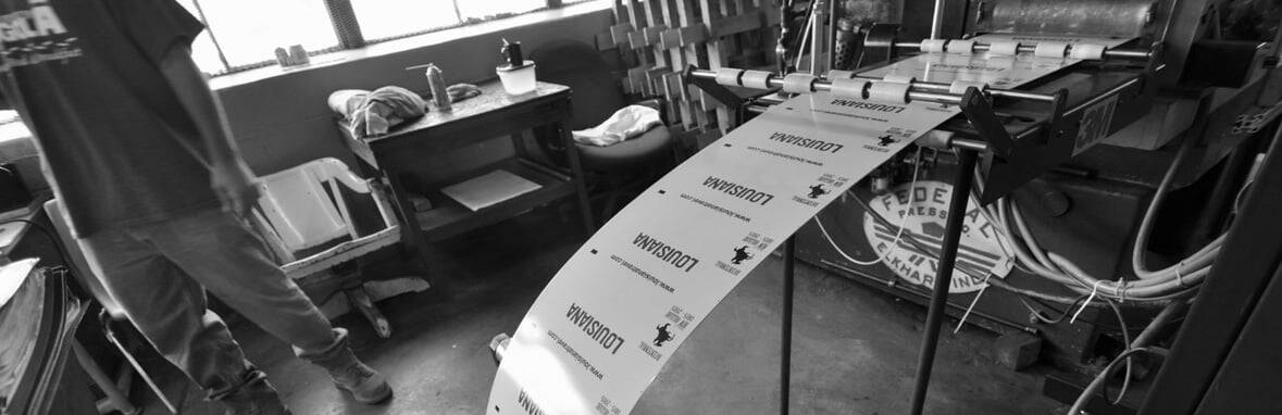 A machine prints labels reading "Ludwina" in a workshop; a person stands nearby with shelves and a table in the background.