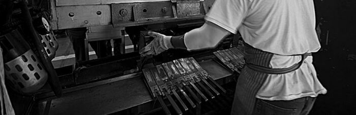 A person wearing gloves and an apron operates a metal press machine, aligning several metal pieces in an industrial setting.