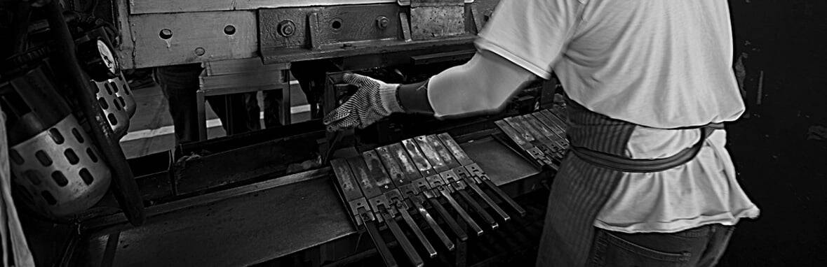 A person wearing gloves and an apron operates a metal press machine, aligning several metal pieces in an industrial setting.