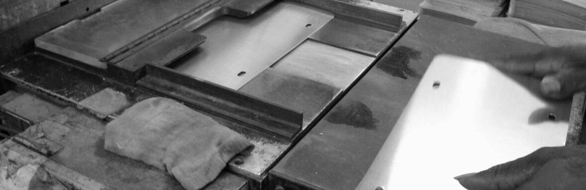 A person’s hands work with metal sheets on an industrial table beside a cloth in a black and white image.