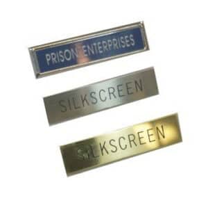 Three rectangular metal name tags in gold or silver: "PRISON ENTERPRISES" on top, two labeled "SILKSCREEN.
