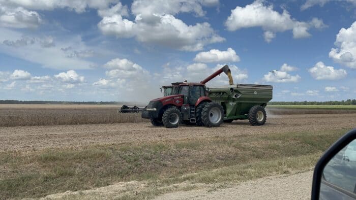 A red tractor pulls a green grain cart in a harvested field under a partly cloudy sky.