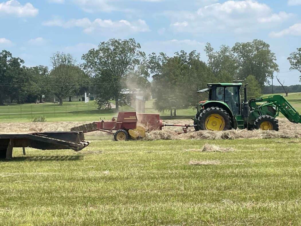 A green tractor pulls a red baler across a field, forming hay bales on a clear day with scattered clouds.