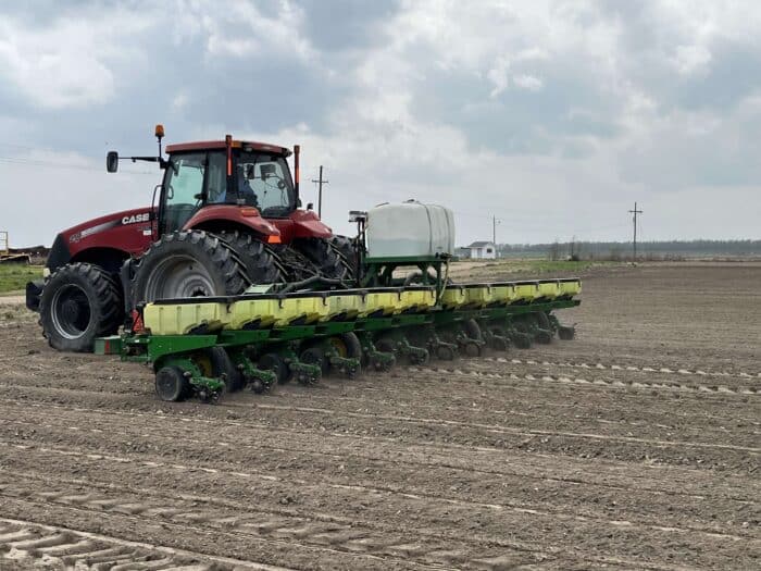A red tractor pulls a multi-row planter across a plowed field, creating rows in the soil under a cloudy sky.
