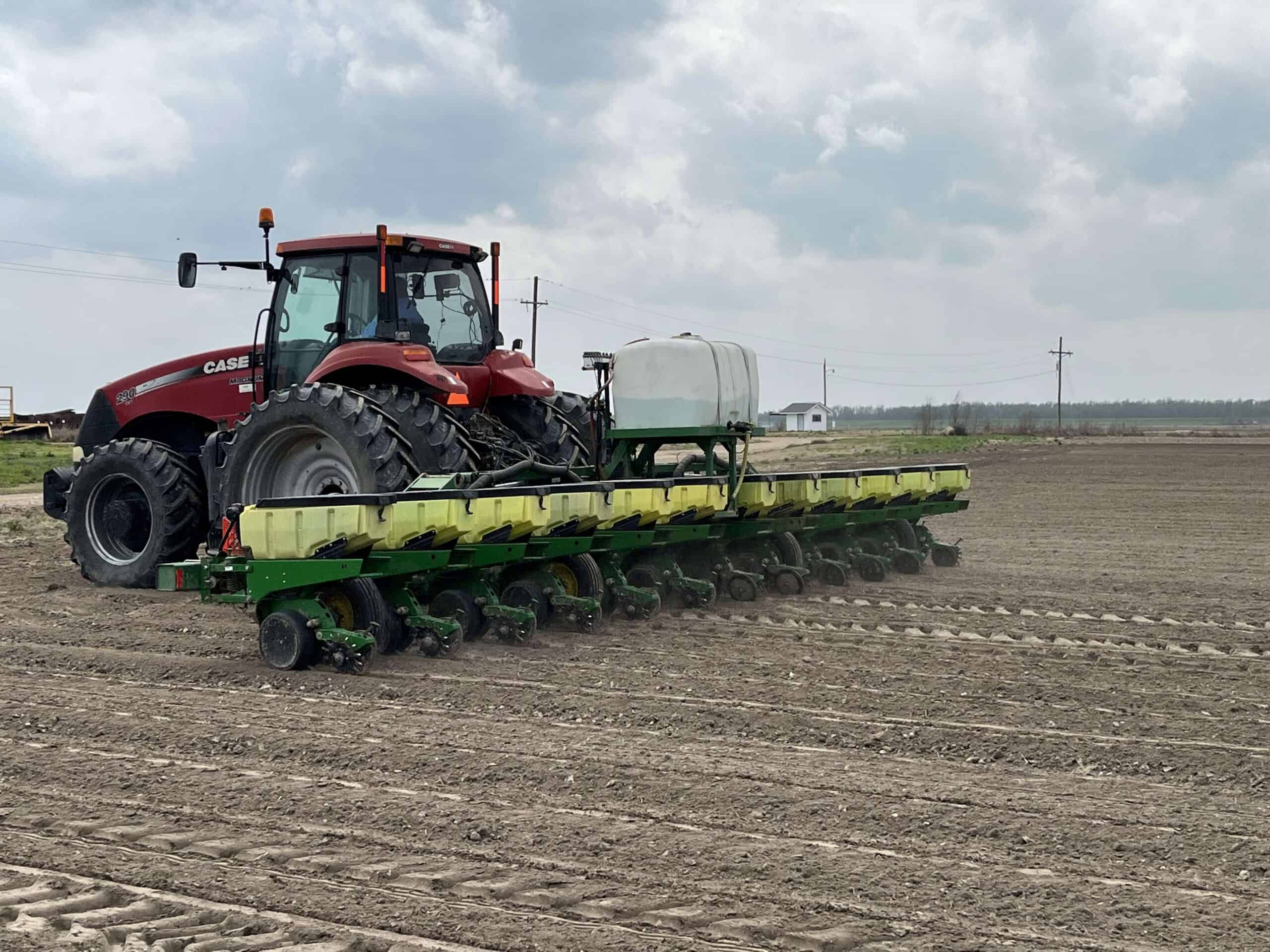 A red tractor pulls a multi-row planter across a plowed field, creating rows in the soil under a cloudy sky.